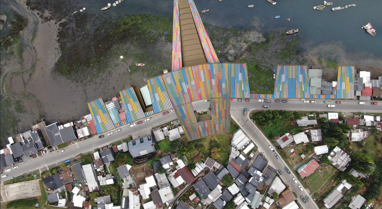 Aerial view of a colorful urban waterfront structure bridging a road and built environment with a wetland or estuarine area. The architectural complex has multicolored rooftops in vertical stripes of red, blue, yellow, and white, resembling corrugated material. The complex spans both sides of the street and includes angled forms that stretch toward the water. Surrounding the site are small residential houses, narrow streets, and patches of green space. Several boats are docked or floating in the nearby water.